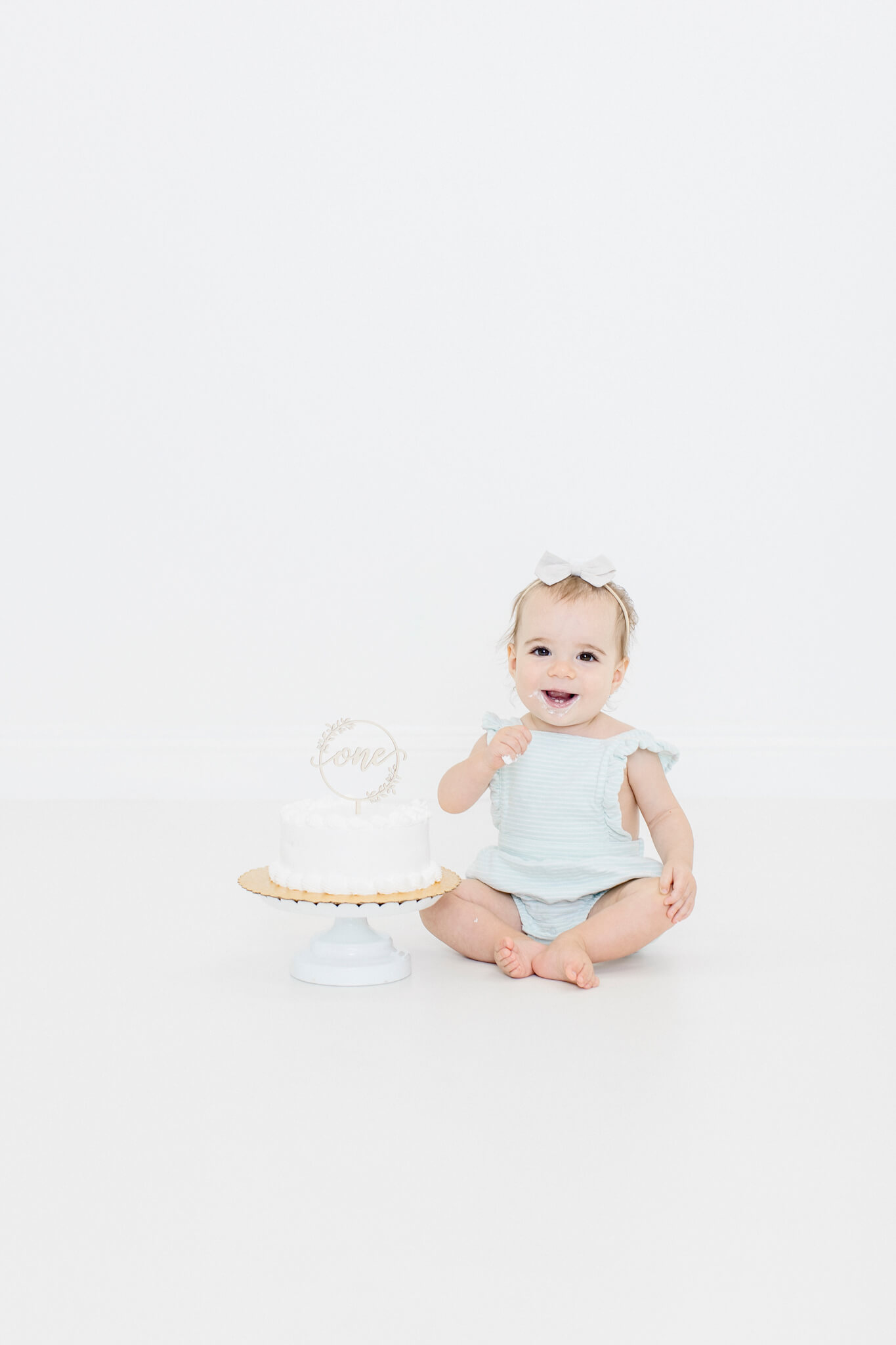 Baby girl sitting on the floor next to a cake on a stand, smiling and waving.