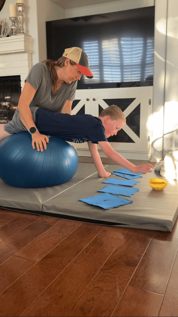 Person on blue exercise ball reaching for blue papers on gray mat, with a child on hands and knees.