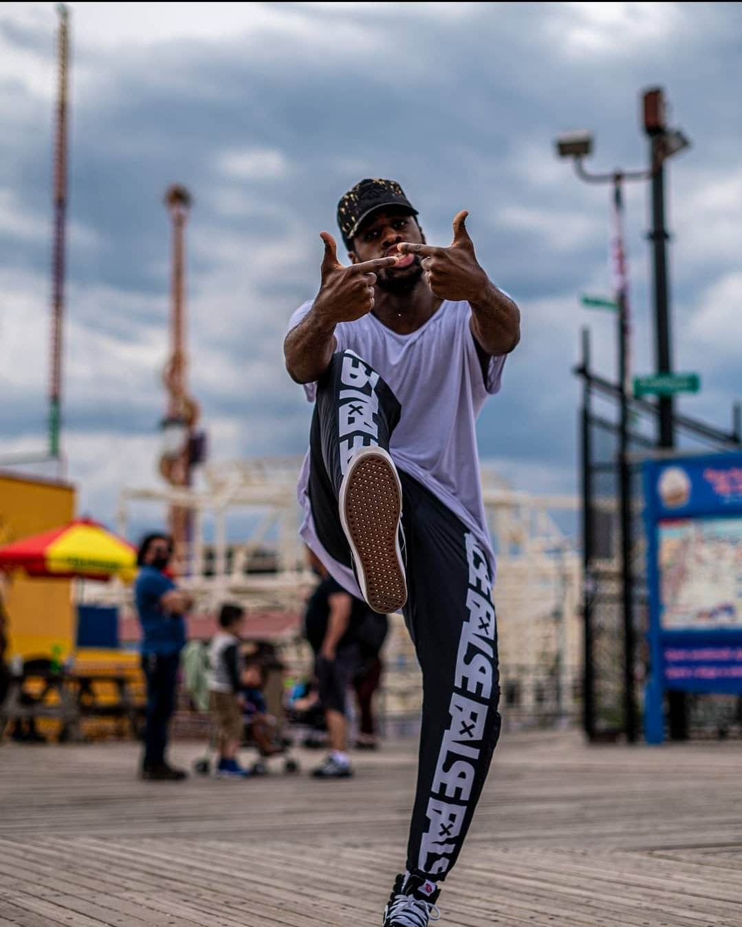 Person performing a high kick on a boardwalk with people and amusement park rides in background.