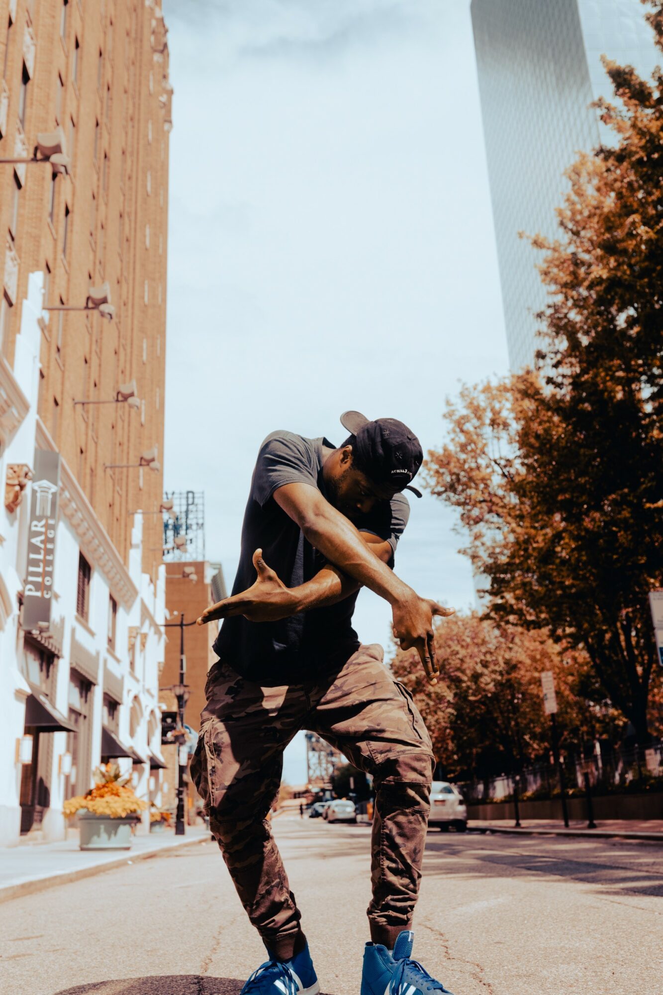 Person dancing outdoors on city street with tall buildings and trees, wearing dark clothing and a cap.