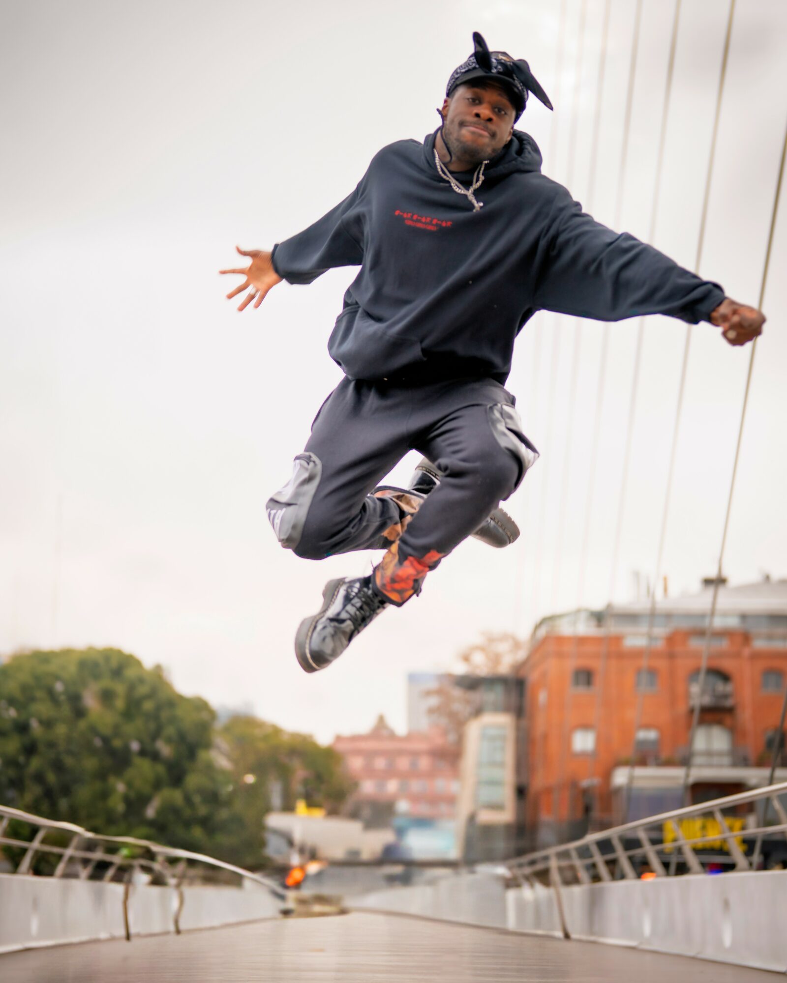 Person mid-air jumping on bridge with city buildings and trees in background.