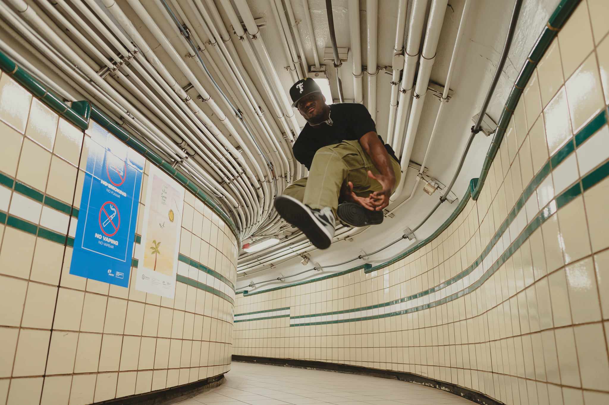 Person walking in a curved tiled corridor holding a bag, pipes on ceiling, no other people visible.