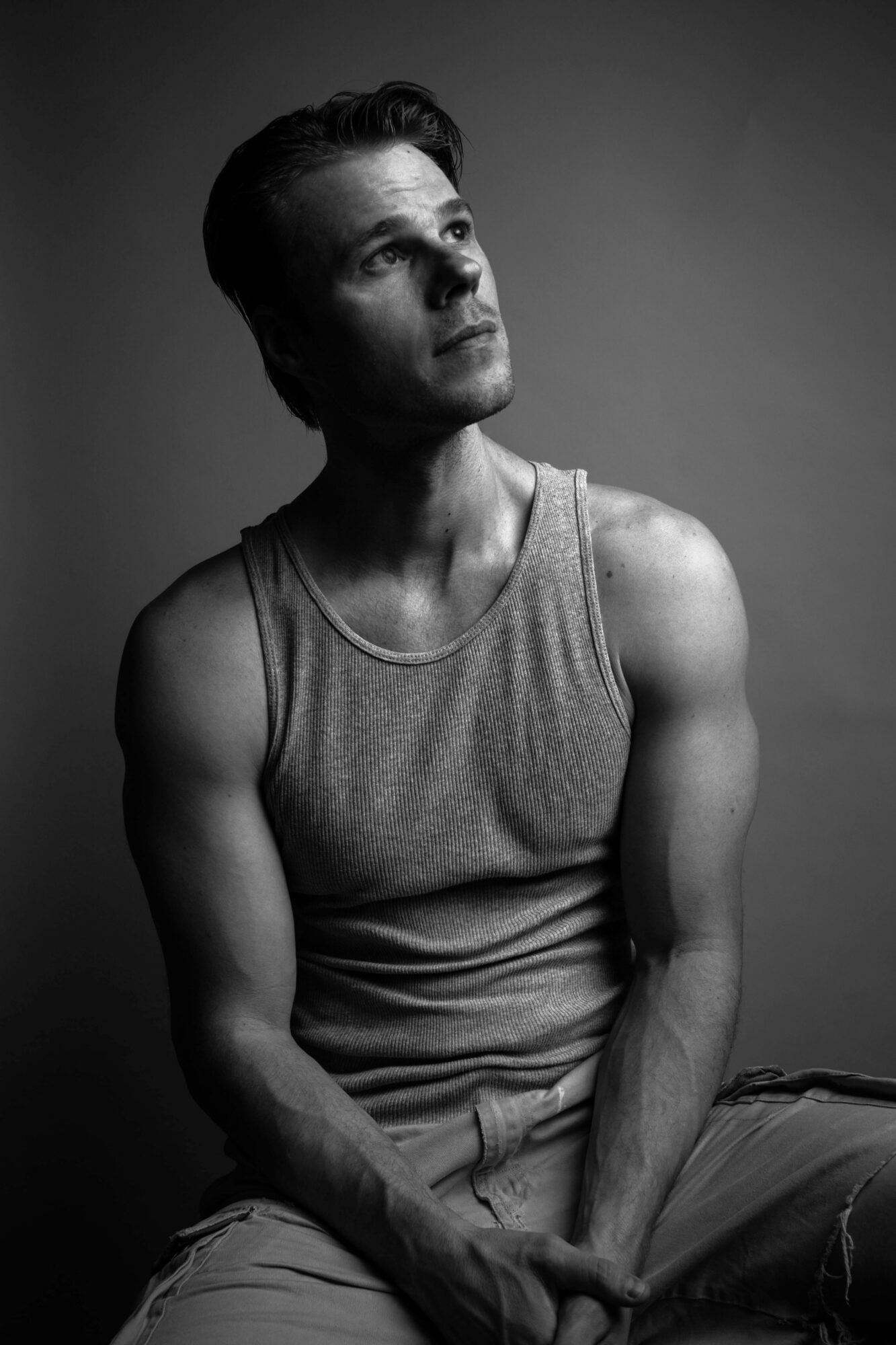 Young man with short hair and sleeveless shirt sitting against a plain background, looking upward.