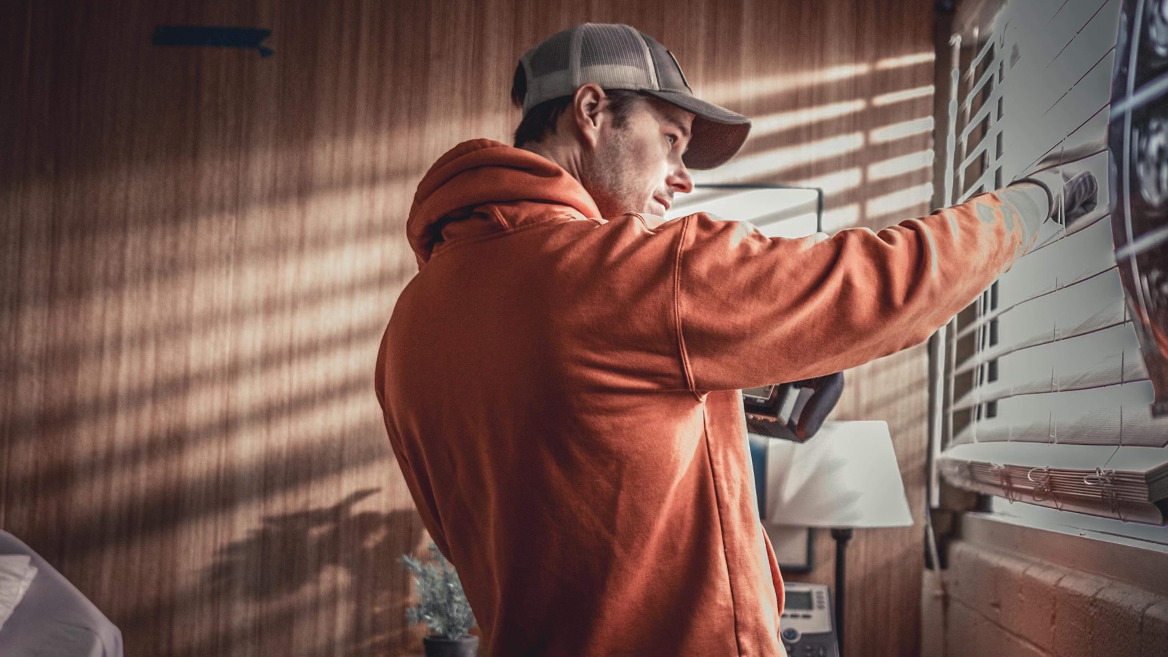 Person in orange hoodie and cap reaching for an object on a wall with blinds casting shadows.