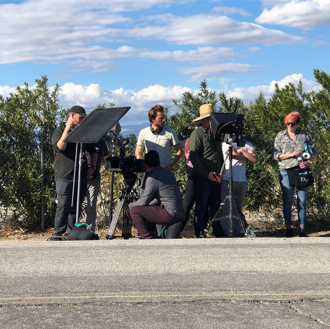 Group of five people filming or photographing outdoors near a fence with trees and blue sky.