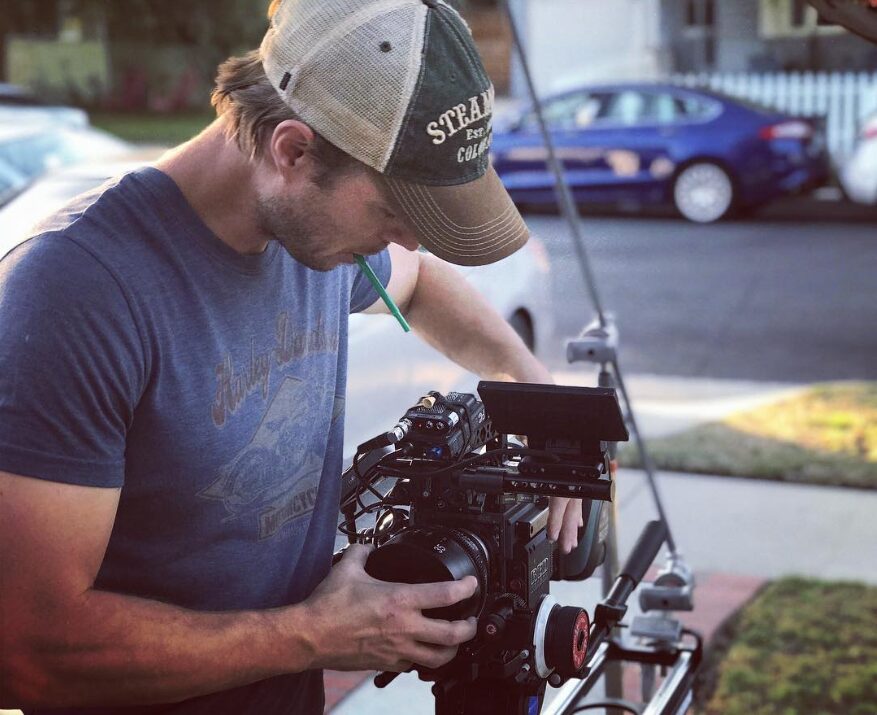 Man wearing a cap and blue shirt operating a camera outdoors on a sunny day.
