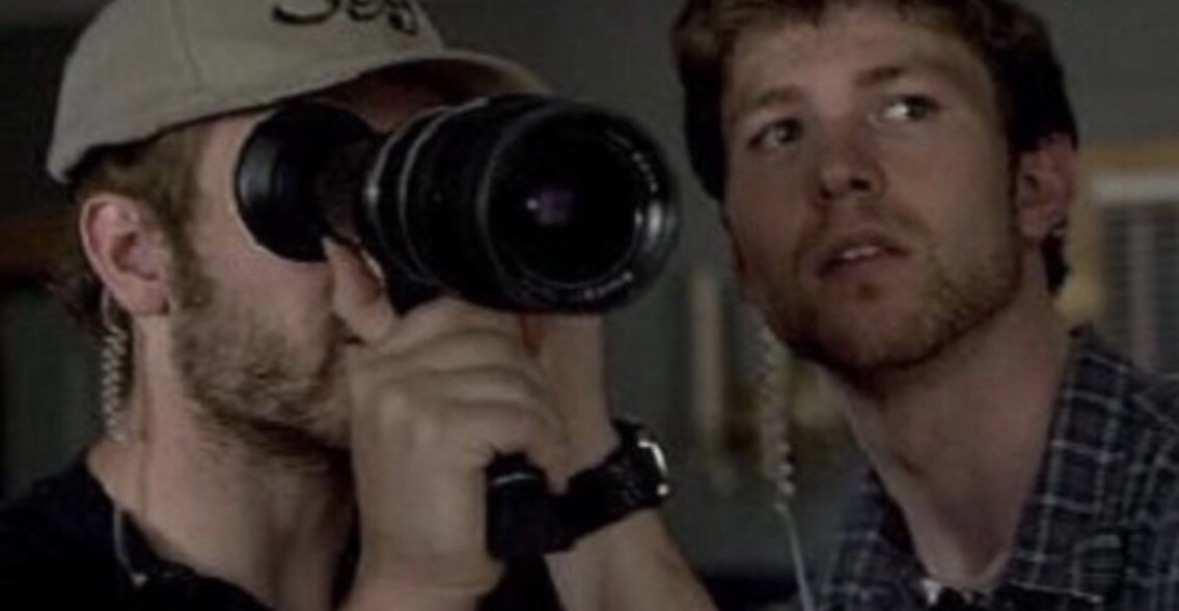 Two men, one with a camera, the other looking at the camera, in an indoor setting.