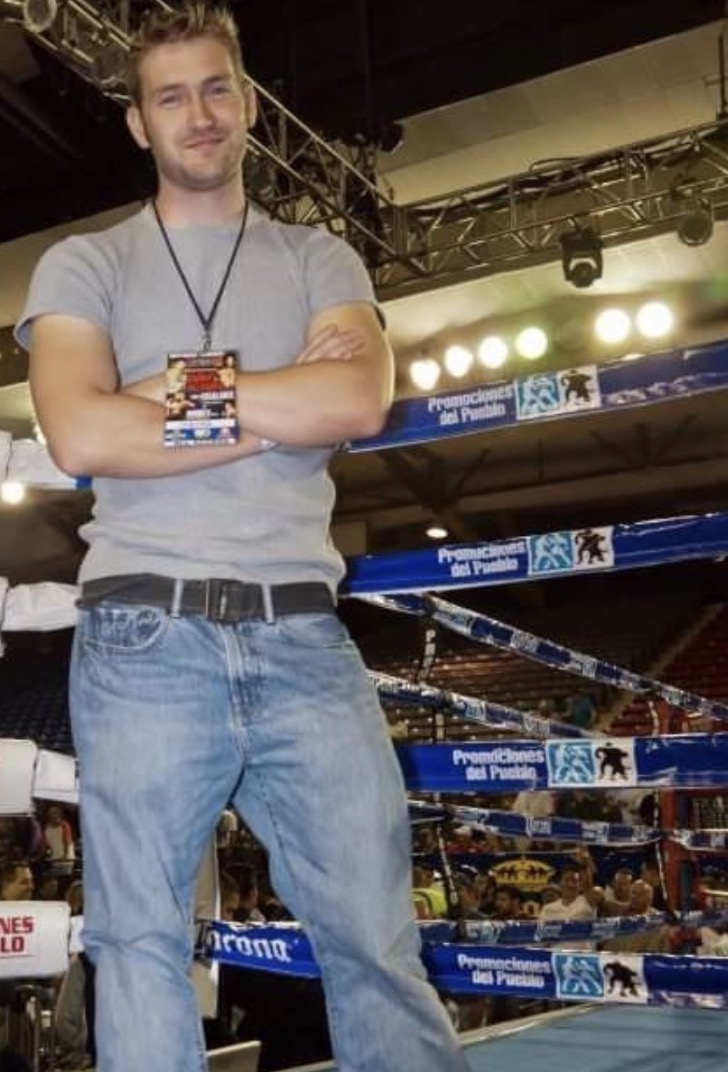 Young man with arms crossed, standing in a boxing ring, wearing a gray t-shirt and jeans, smiling.