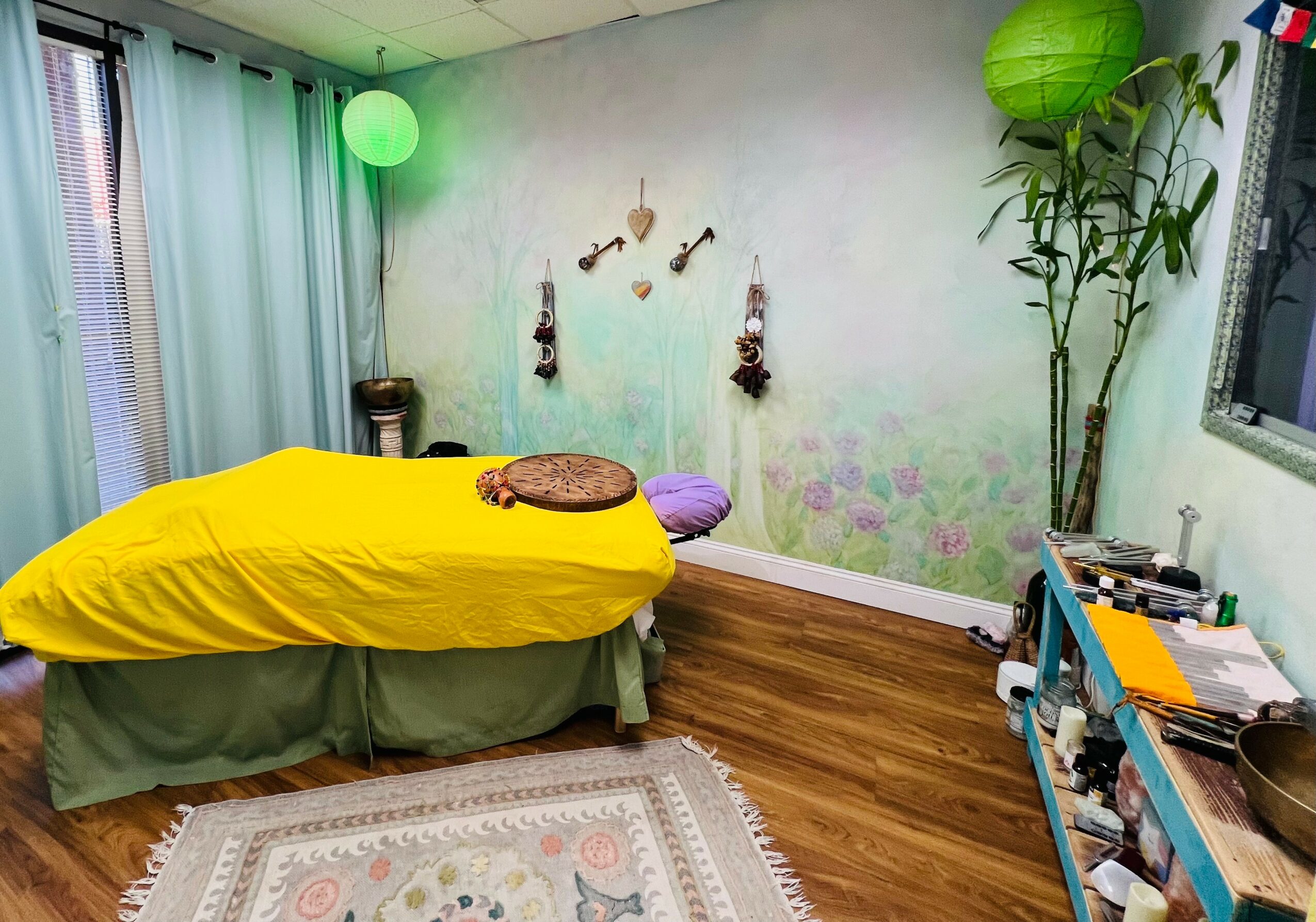 Bedroom with bed covered in yellow blanket, green bed skirt, purple pillow, and wall decorations, wooden floor, and a tall plant.