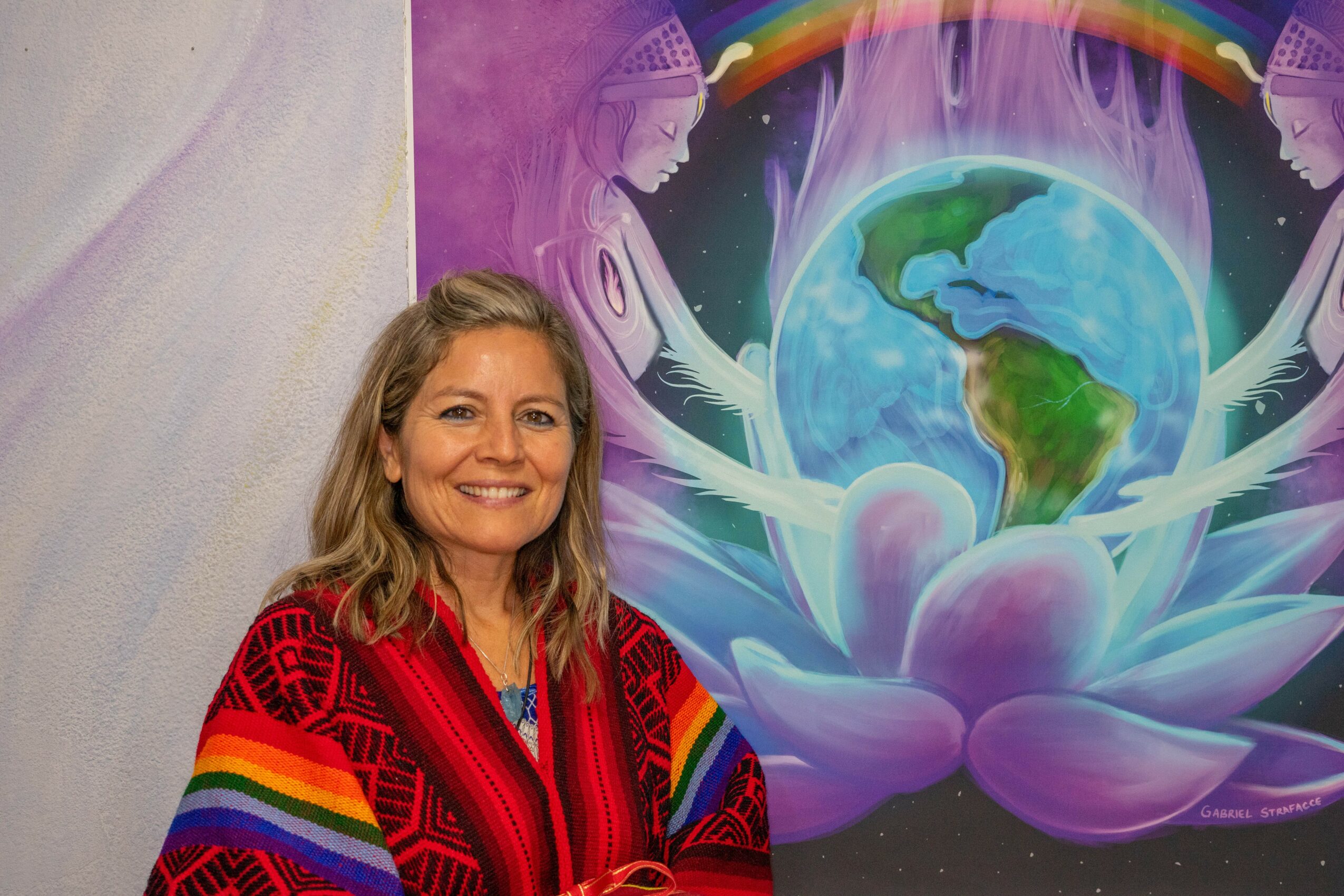 Woman with blonde hair smiling, wearing a colorful rainbow-patterned sweater, standing in front of a vibrant mural of Earth and abstract figures.