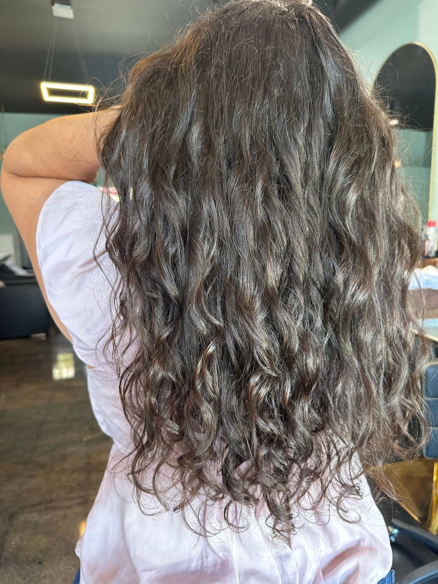 Person with long, wavy hair seen from behind, wearing a white shirt, in an indoor setting.