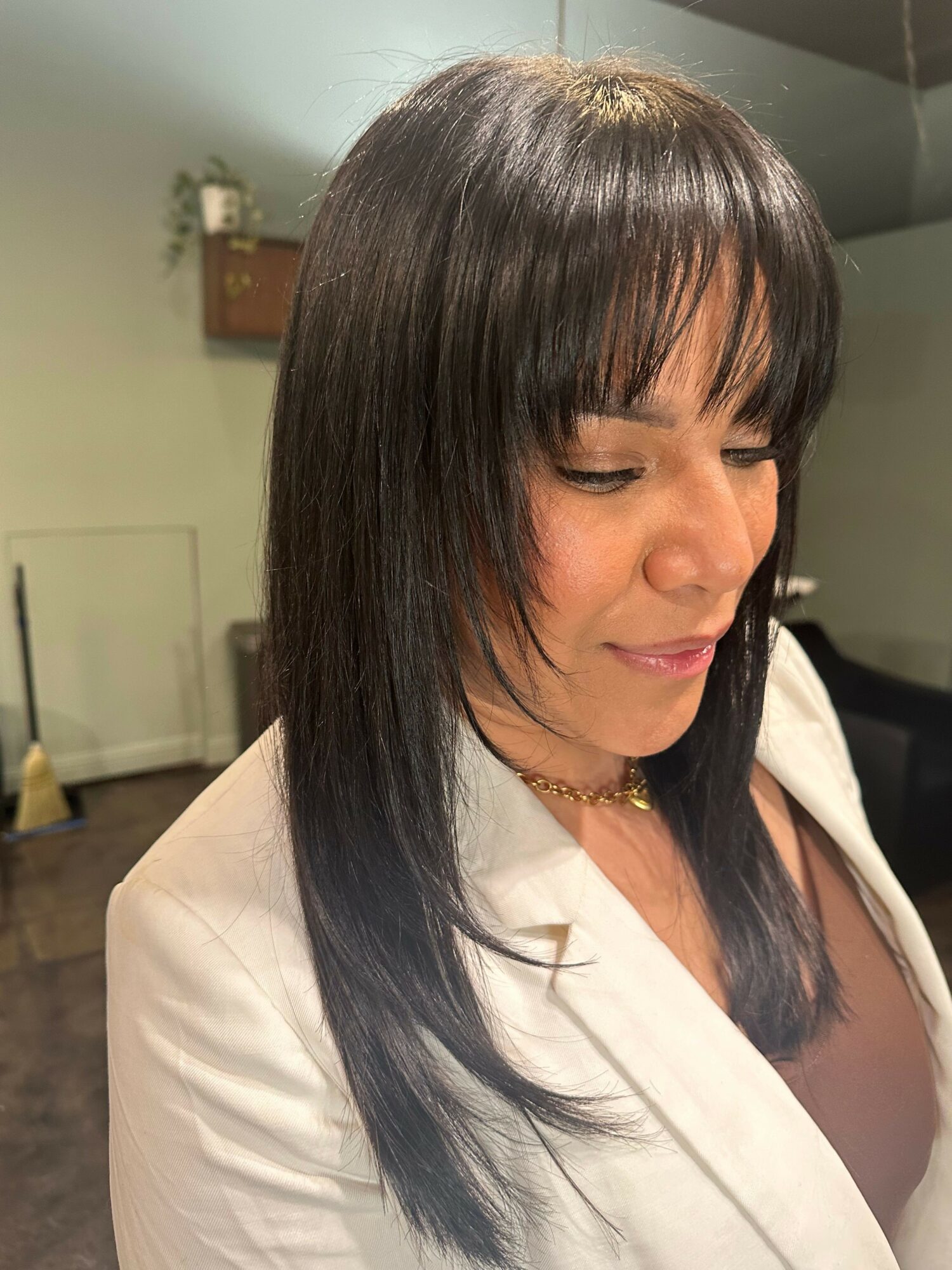Woman with straight, dark hair and bangs, wearing a white blazer and gold necklace, looking down.