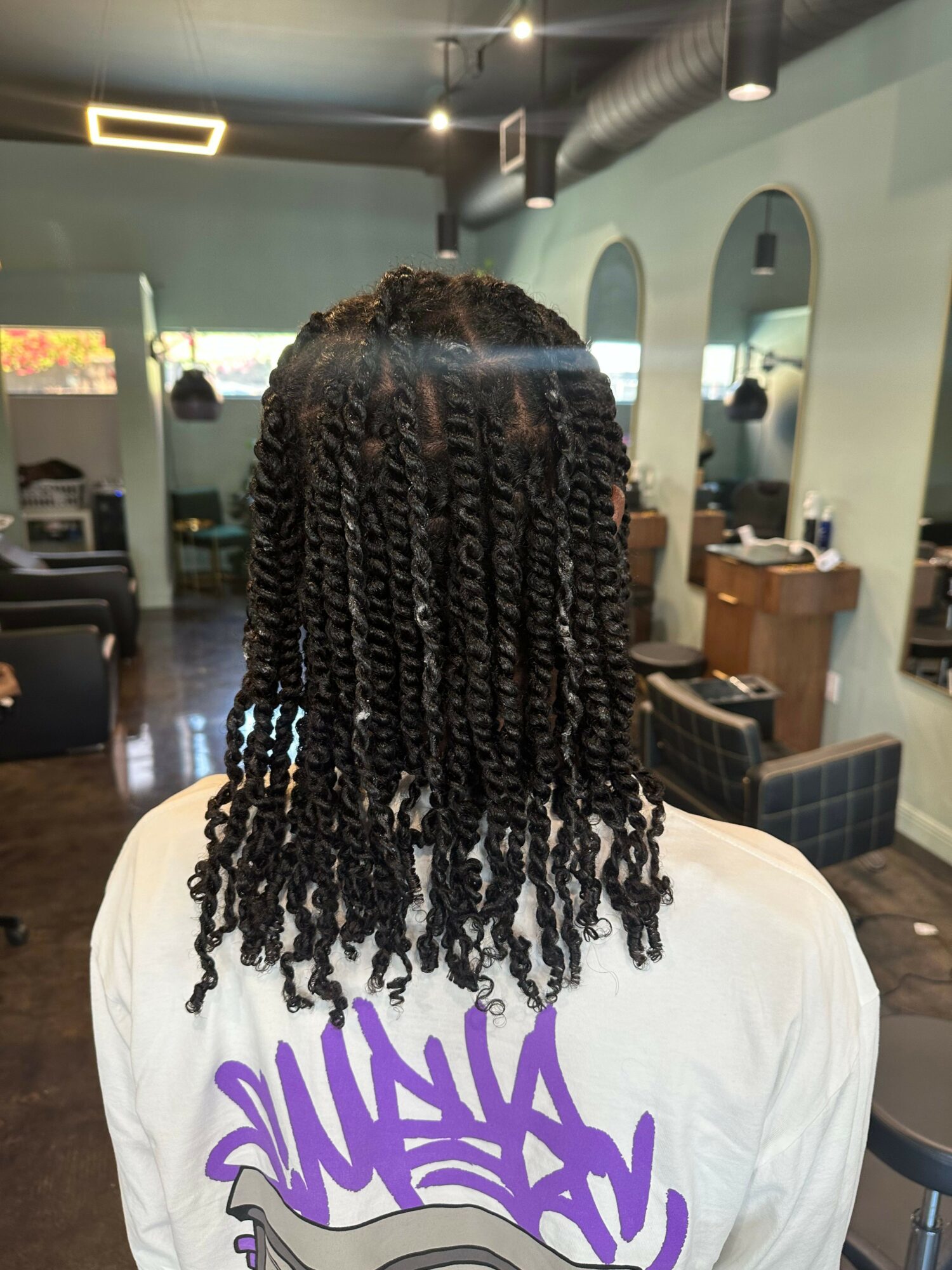 Back of person with curly, twisted hairstyle in a salon setting with mirrors and chairs.