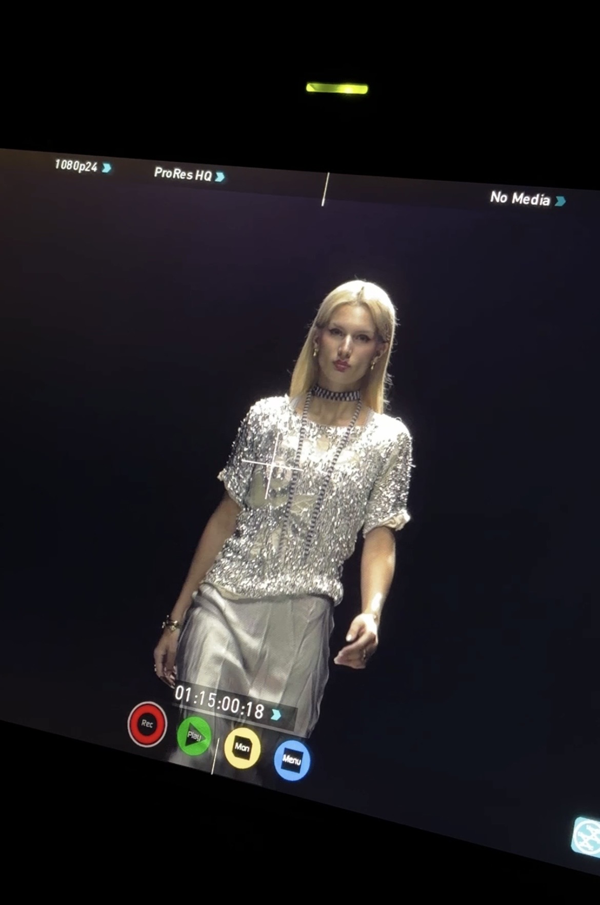 Person with blonde hair wearing a shiny, sequined top and light-colored pants, standing against a dark background.