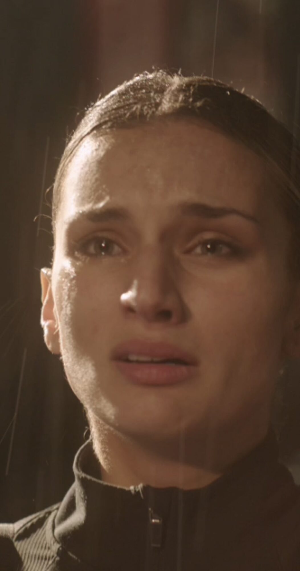 Close-up of a woman with a distressed expression, wet hair, and a dark jacket, indoors with warm lighting.