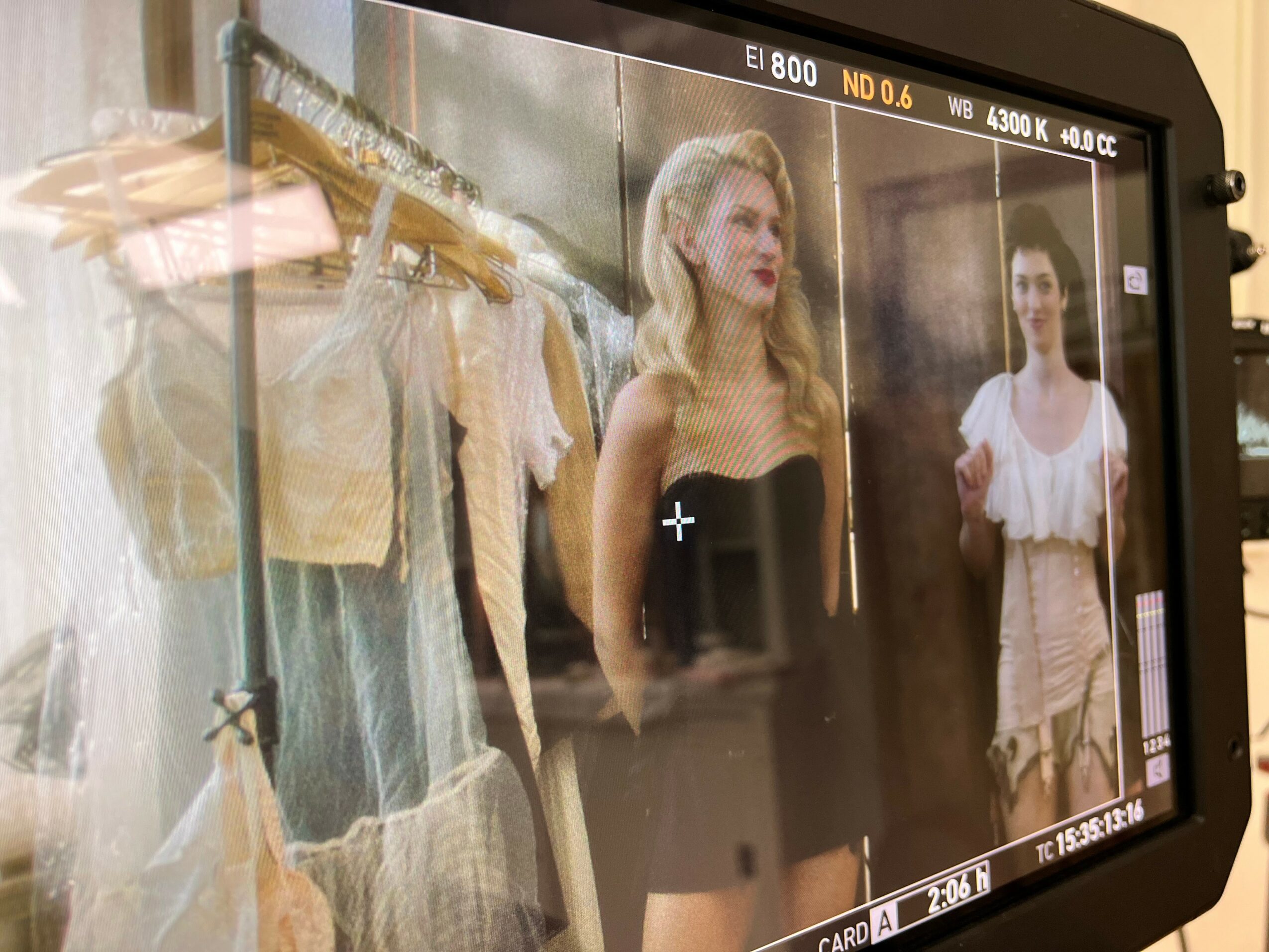 Two women standing in front of a clothing rack with dresses and tops, one in a black dress, the other in a white top.