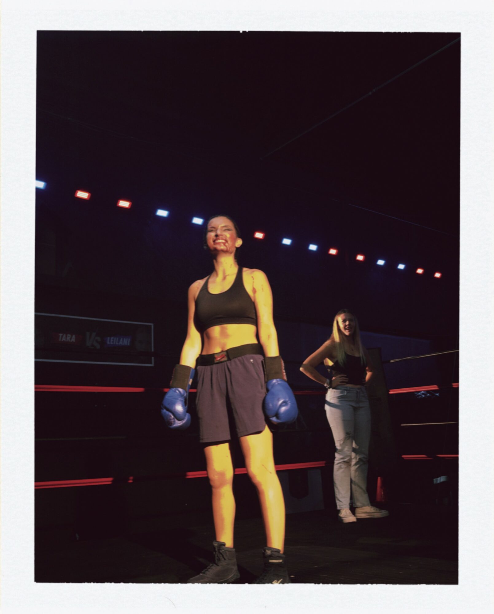 Female boxer in gloves and athletic outfit standing in a boxing ring with a person in the background.