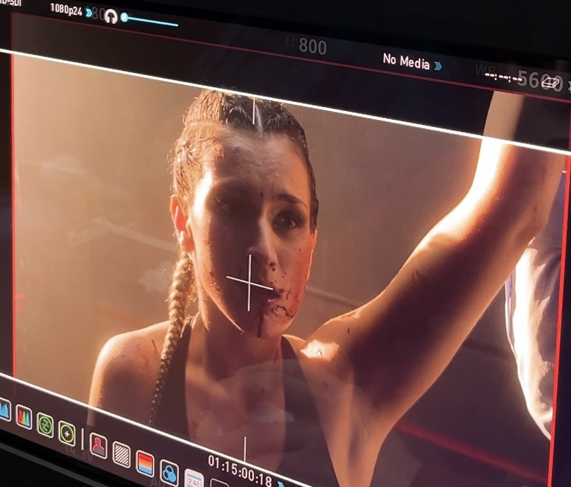 Woman with braided hair and raised arm, looking at camera, on a film set monitor.