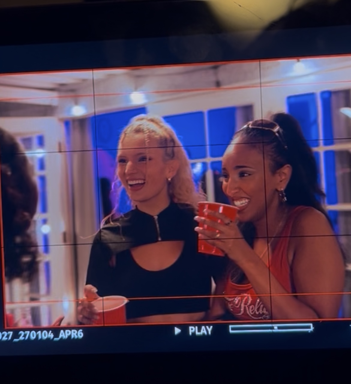 Two women smiling and holding drinks at a social event, with a blue-lit background and large windows.
