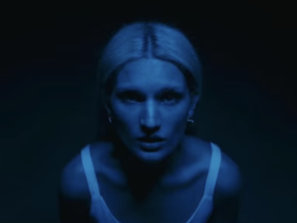 Woman with blonde hair illuminated by blue light, facing forward with a serious expression, dark background.