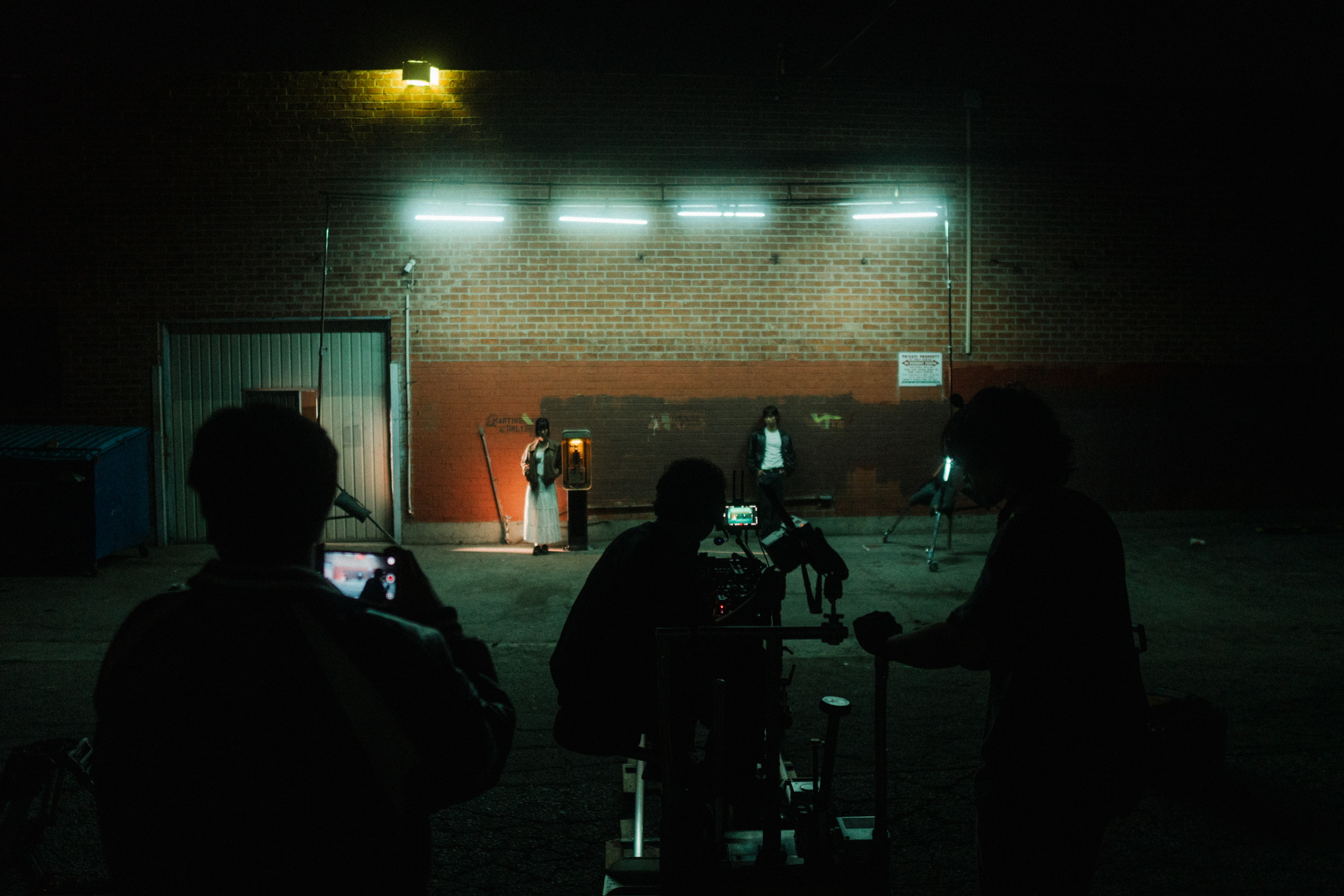Silhouettes of people in a dark outdoor area near a brick wall with bright lights above, some using phones or cameras.