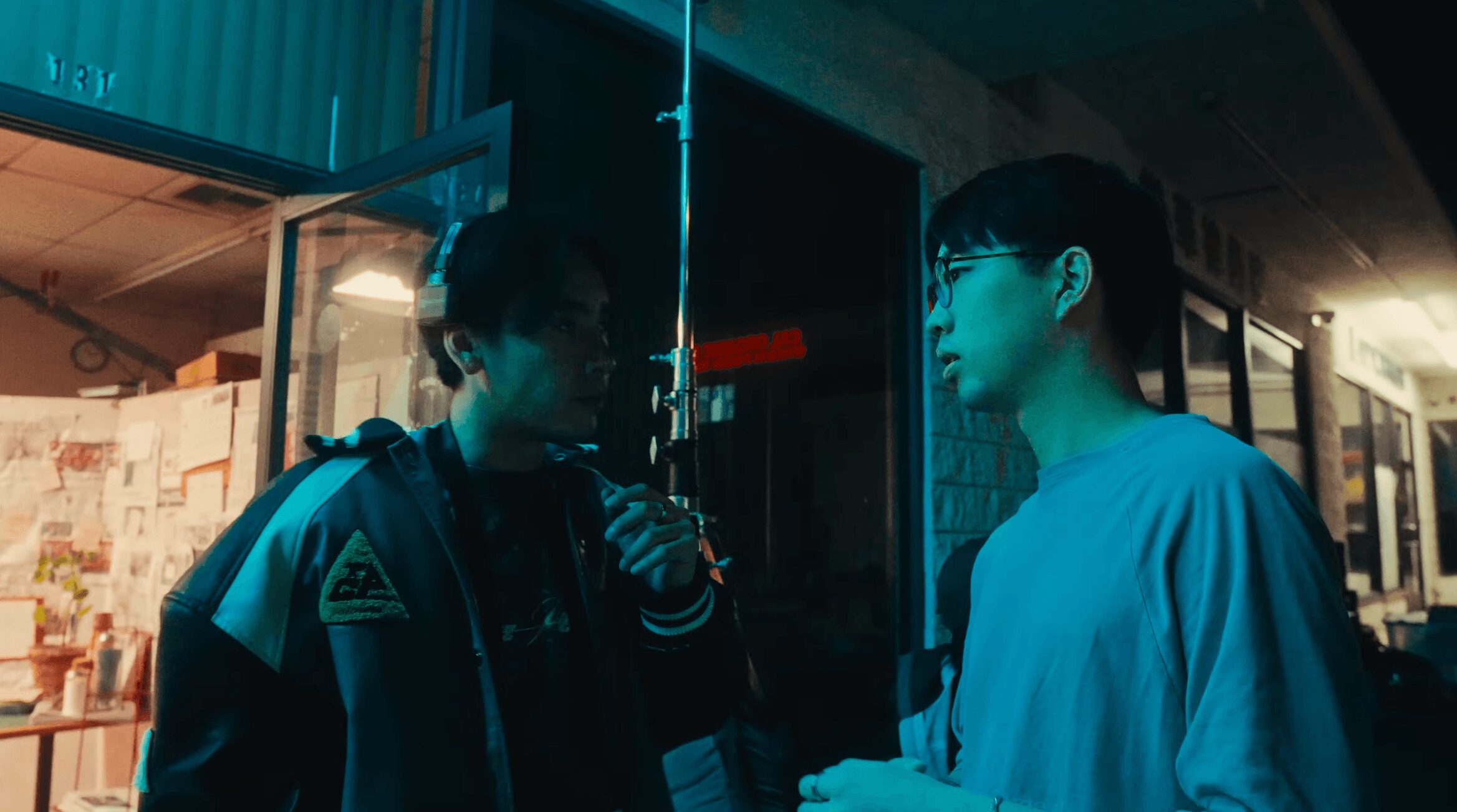 Two young men stand outside a building at night, engaged in conversation, with city lights in the background.