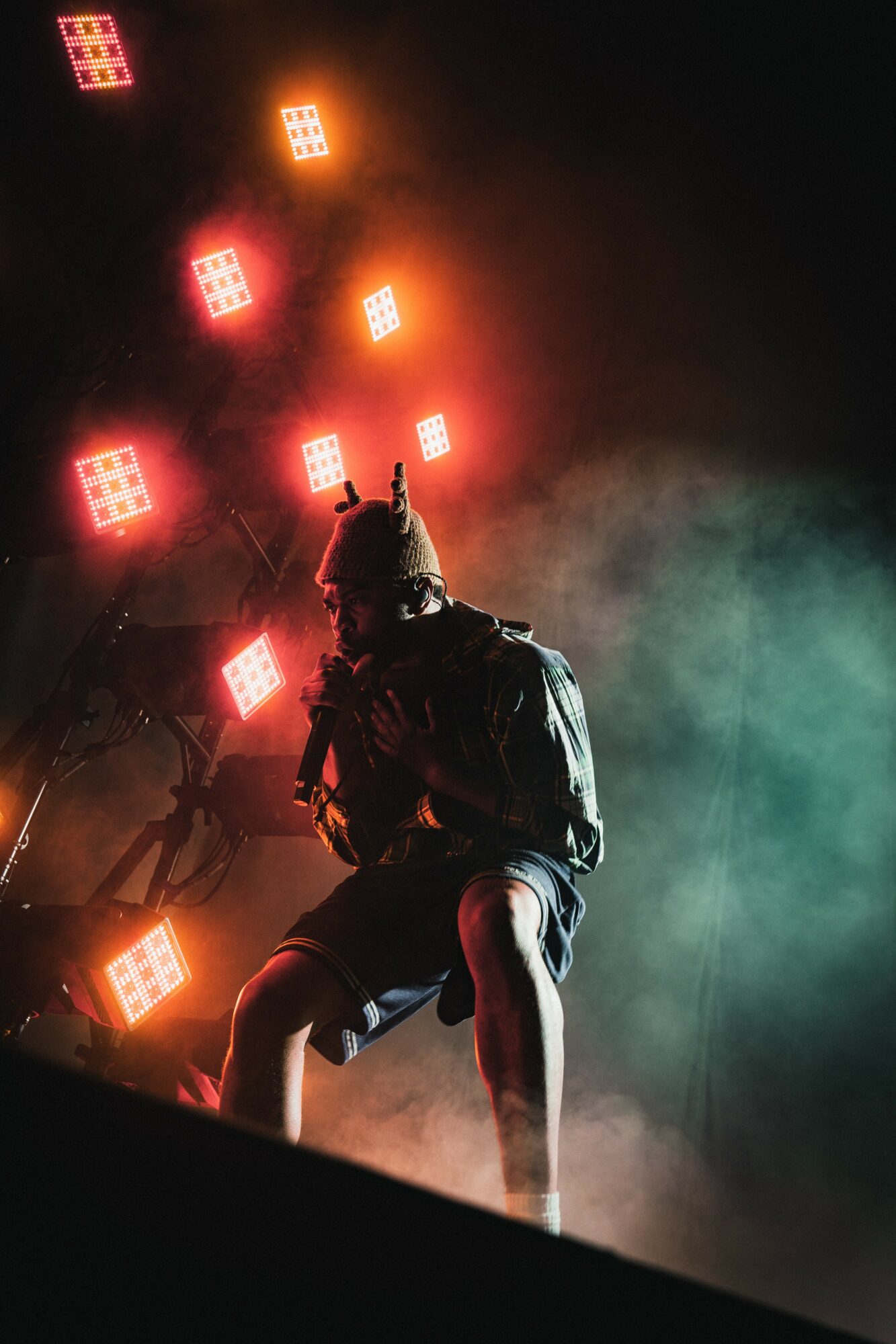 Person wearing a mask and hoodie, sitting on a stage with colorful lights and smoke in the background.