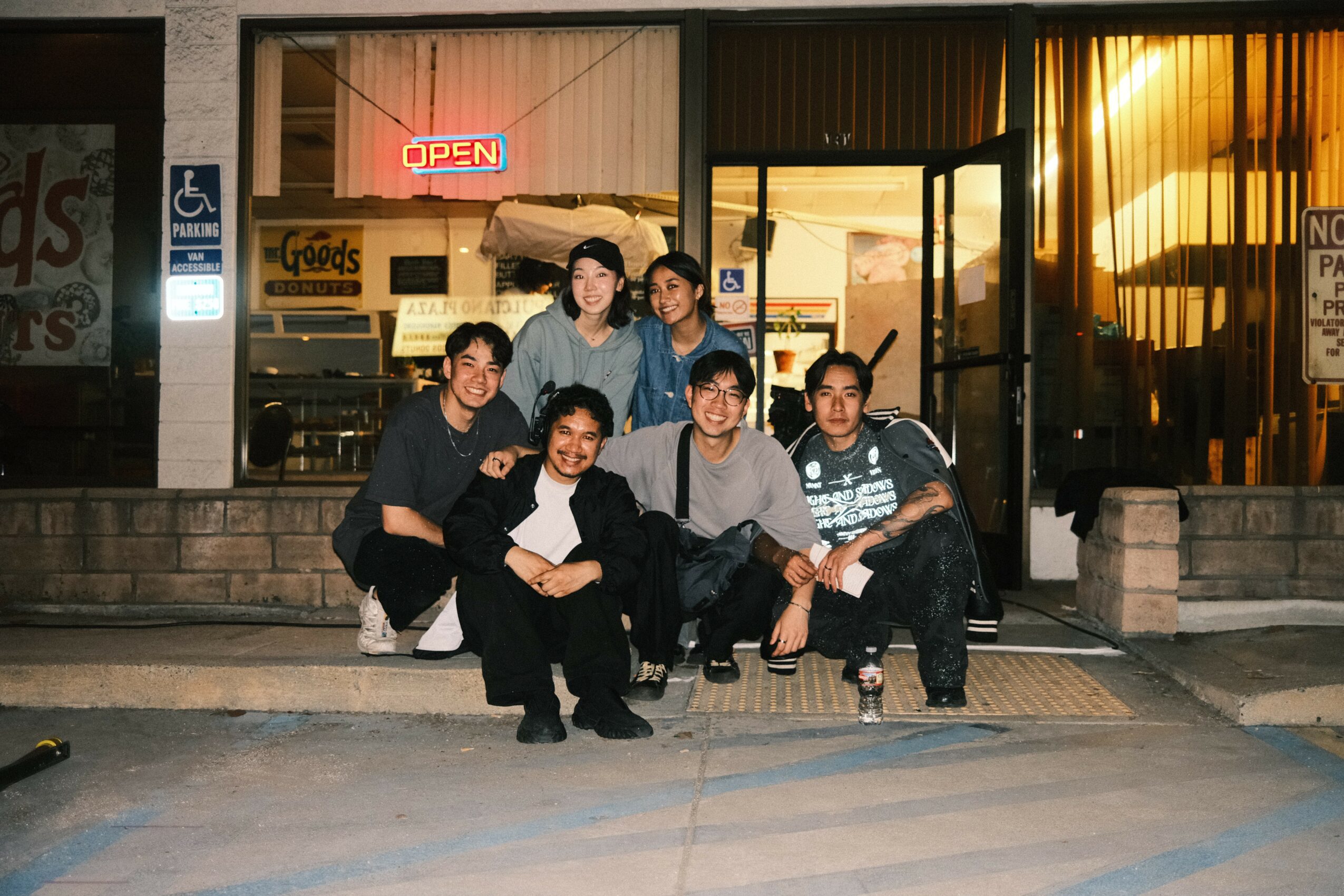 Group of seven people sitting and standing outside a store with an illuminated 'OPEN' sign, during sunset.