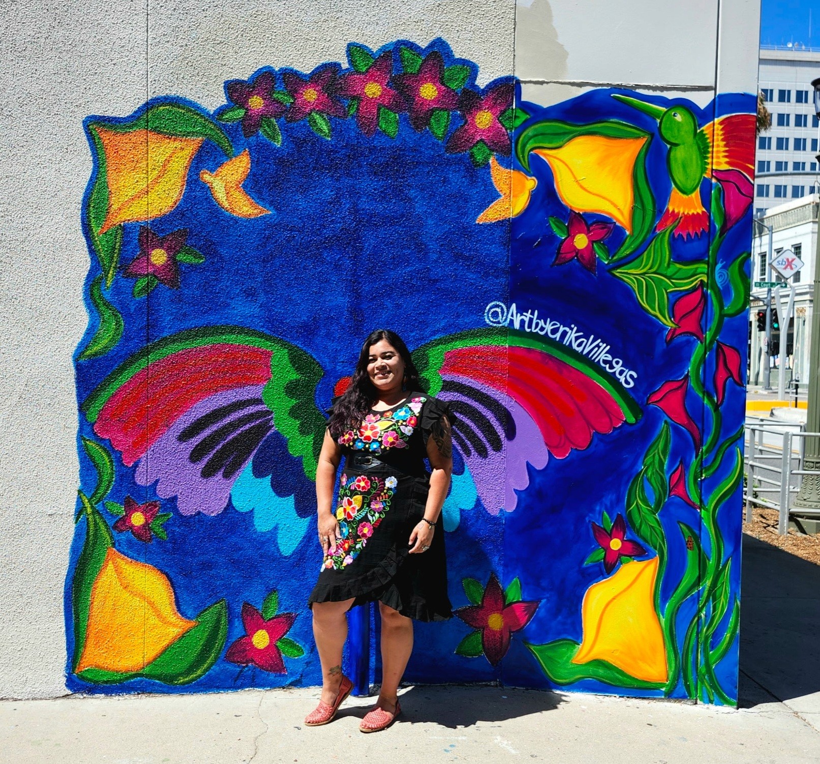 Woman standing in front of colorful butterfly mural with flowers and vibrant patterns on wall.