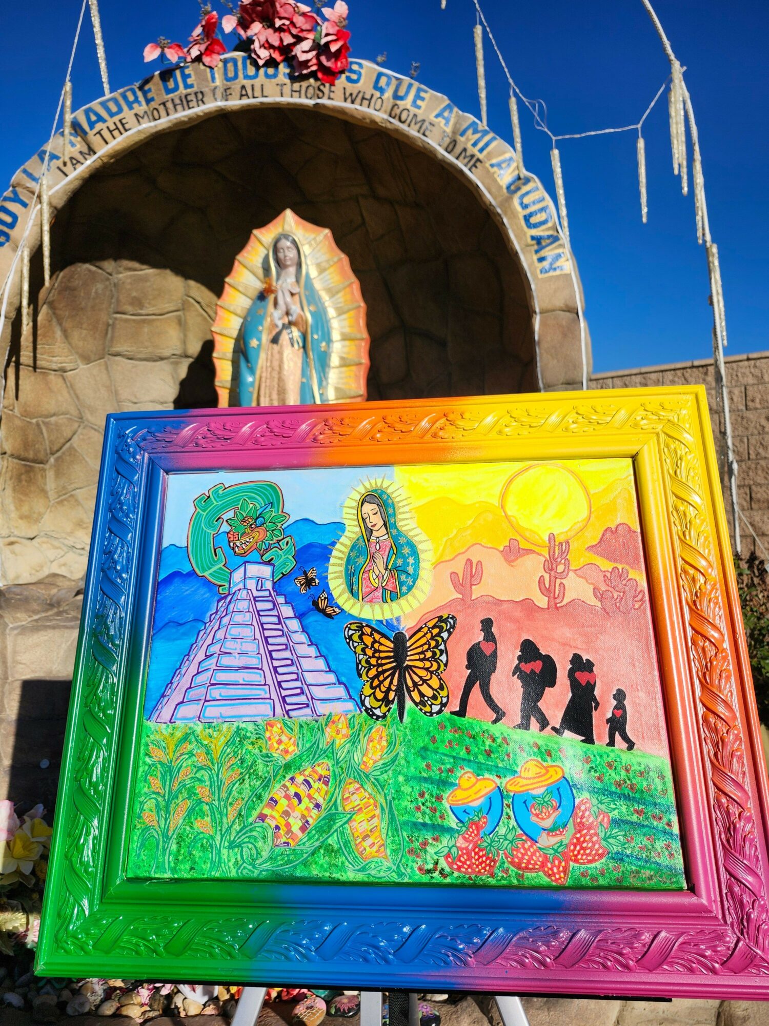 Colorful framed painting of religious figures, butterflies, and nature scenes in front of a grotto with a statue of the Virgin Mary.