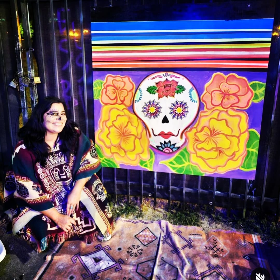 Woman sitting next to a colorful sugar skull painting with flowers and stripes, outdoors at night.