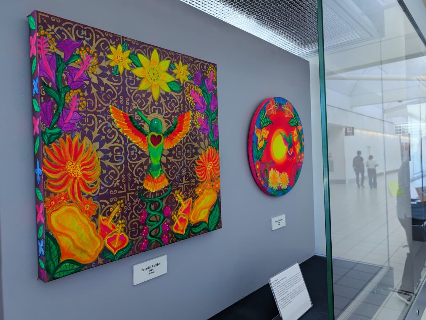 Two colorful, vibrant paintings displayed on a gray wall in an art gallery, with visitors visible outside the glass wall.
