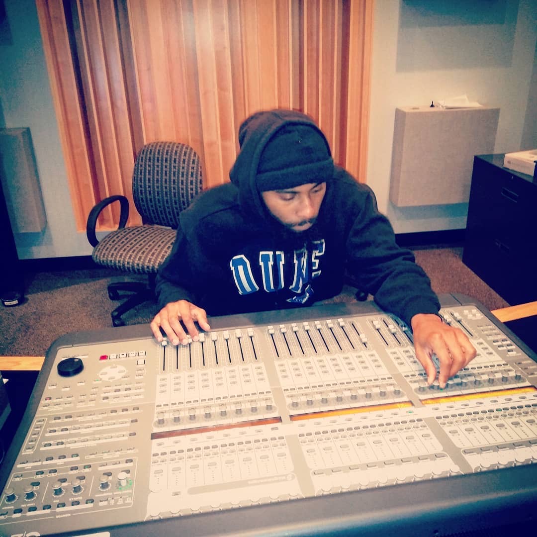 Person wearing a black hoodie and head covering, working at a large audio mixing console in a recording studio.