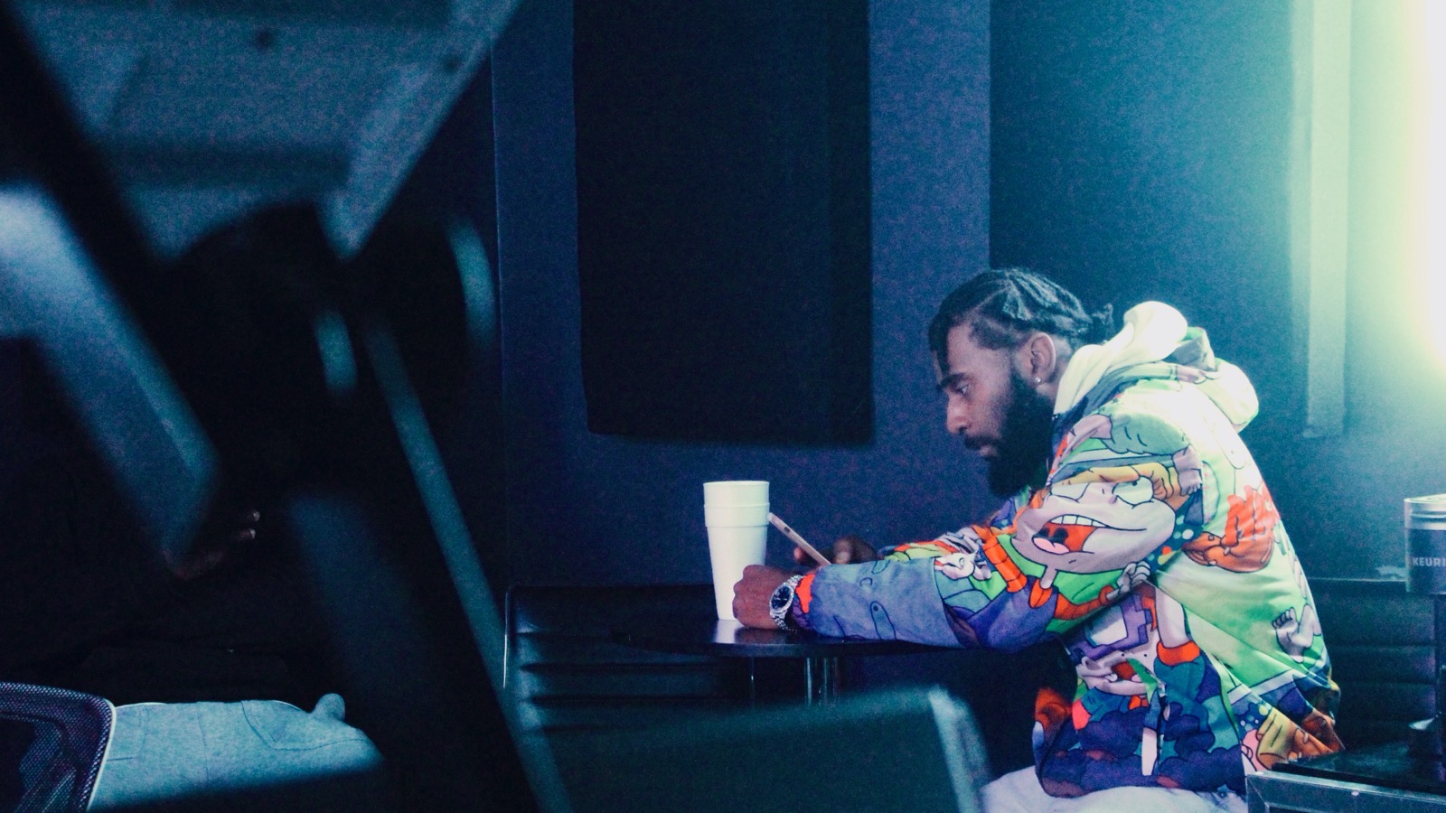 Person with colorful jacket sitting at a table with a drink and a cup, in a dimly lit room.