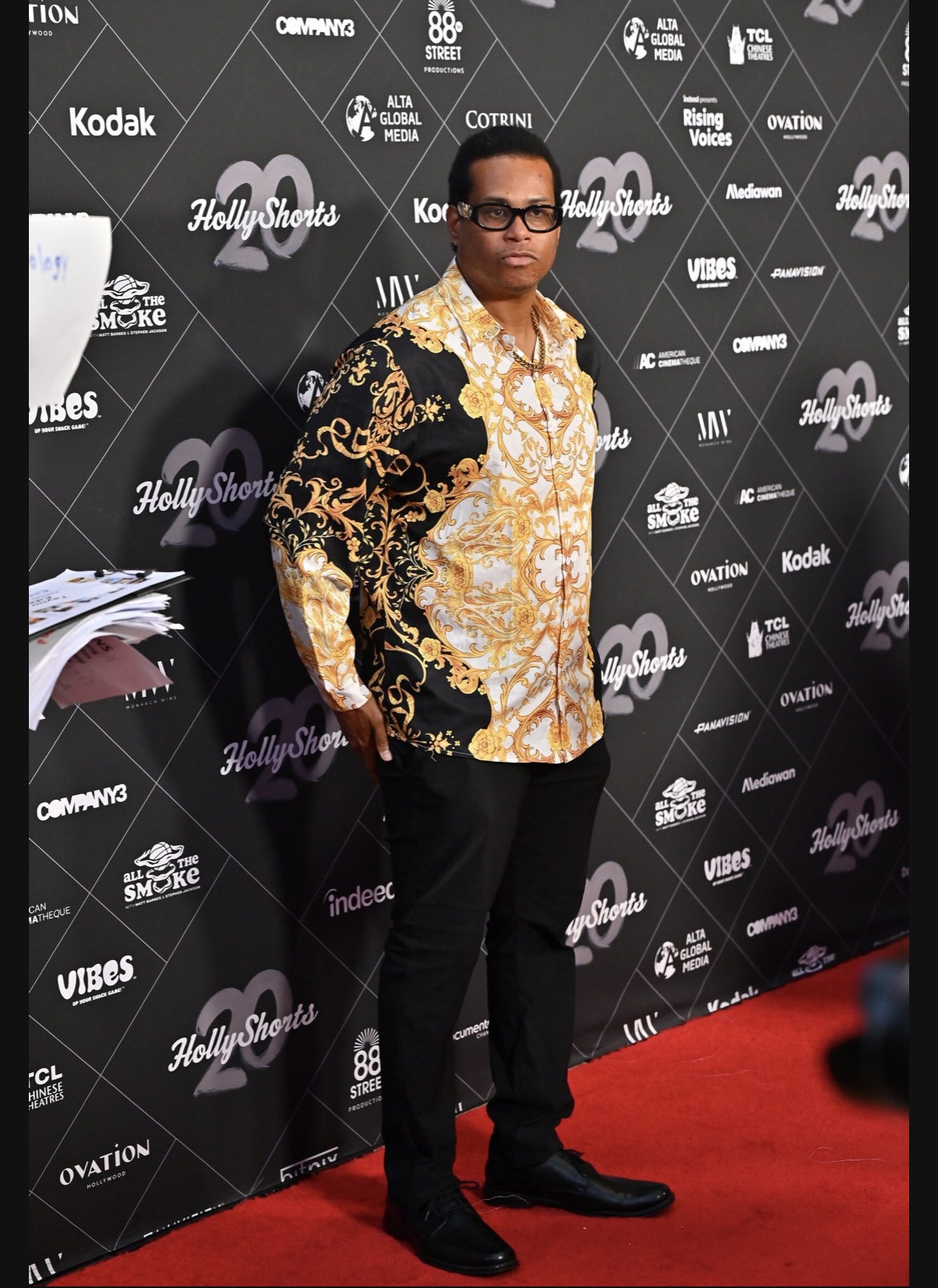 Person standing on red carpet in front of black backdrop with logos, wearing glasses, patterned shirt, and black pants.