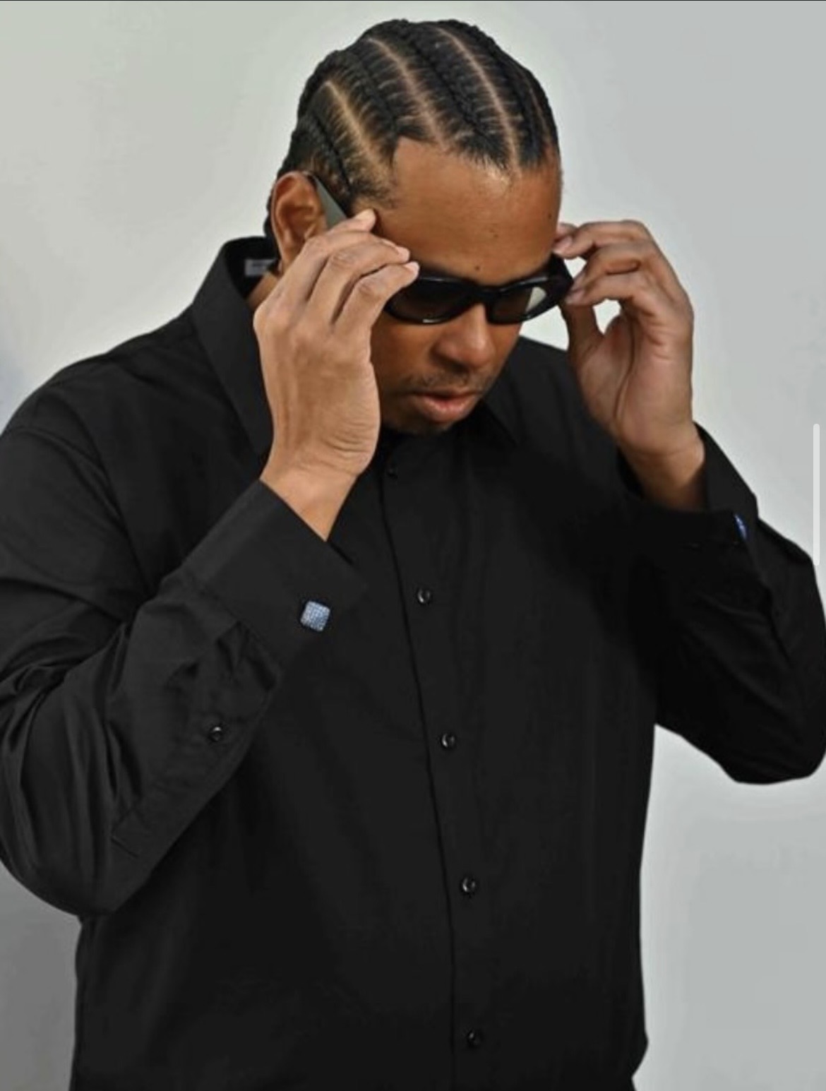 Person with braided hair adjusting sunglasses, wearing a black shirt, against a plain background.