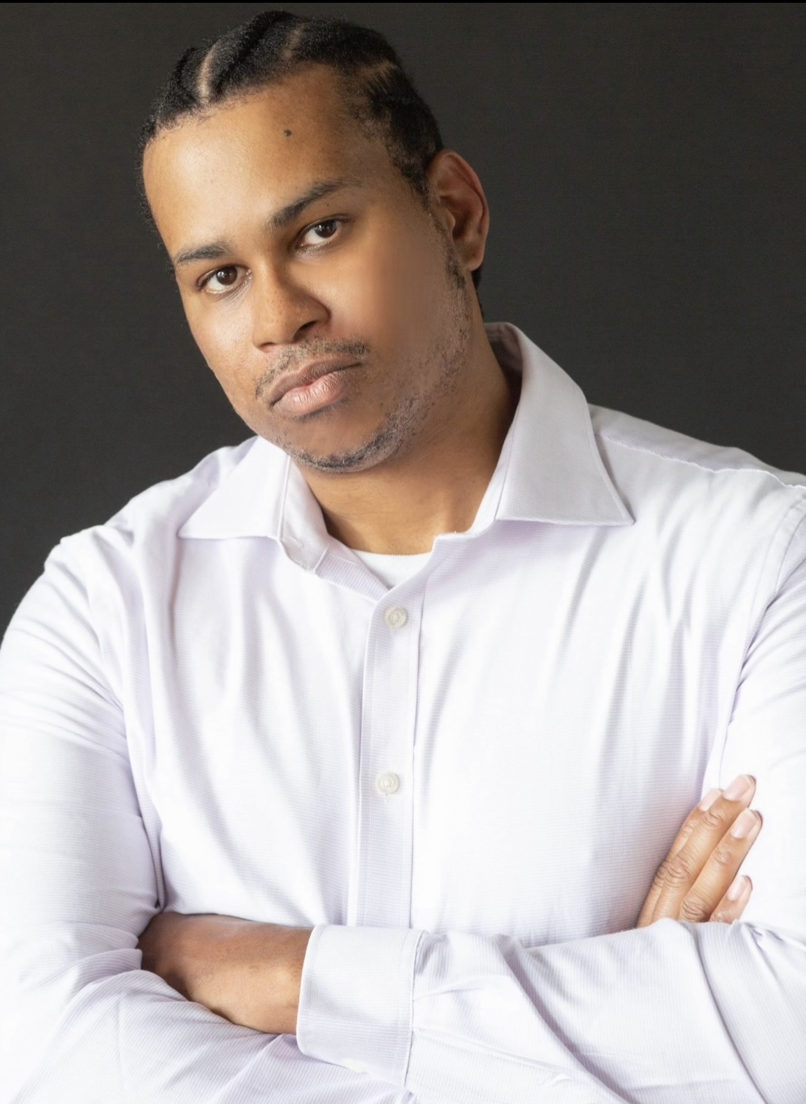 Man with braided hair wearing a white shirt, arms crossed, looking at camera against dark background.