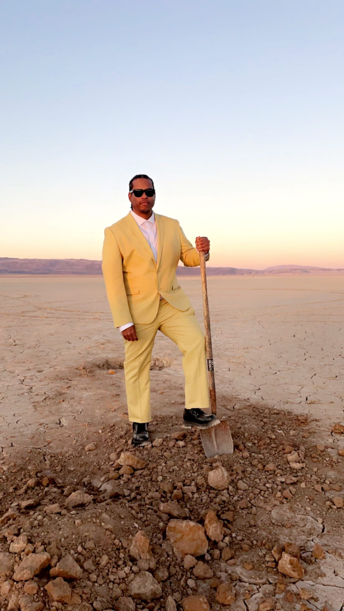 Man in yellow suit holding a shovel in a barren landscape during sunset.