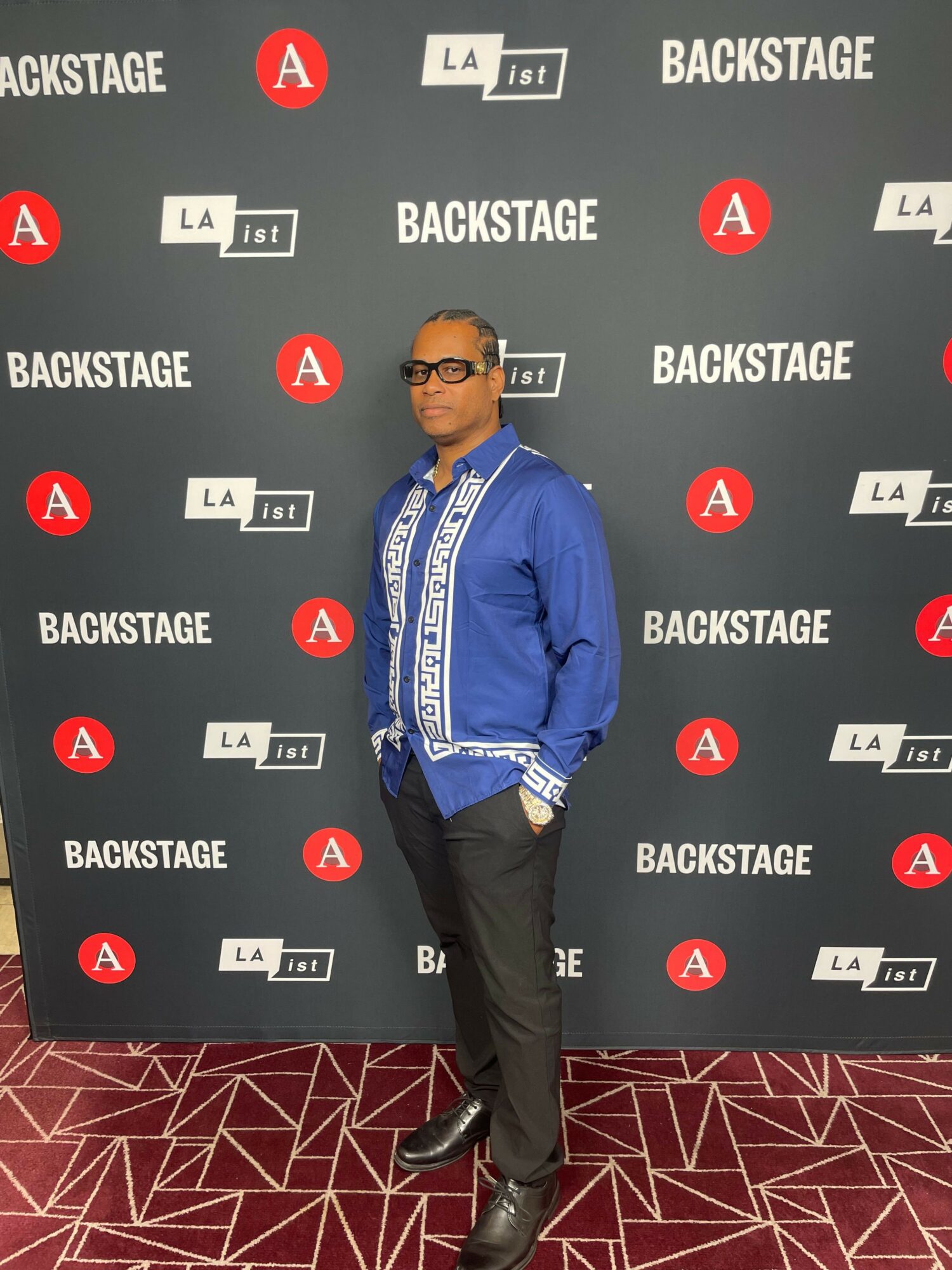 Man in blue shirt and black pants standing in front of a backdrop with logos and text.