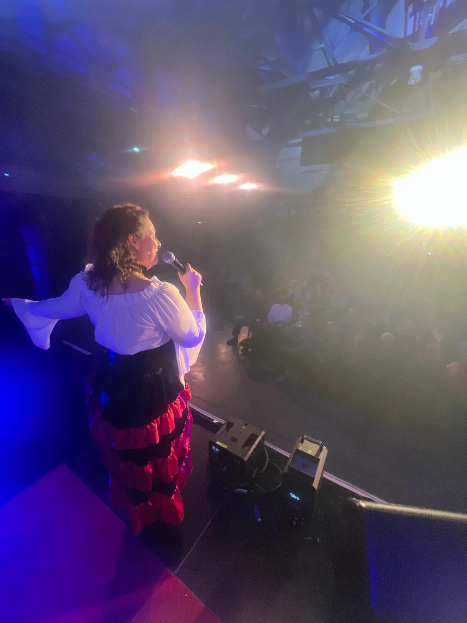 Performer with curly hair singing into a microphone on stage with bright lights and fog effects.