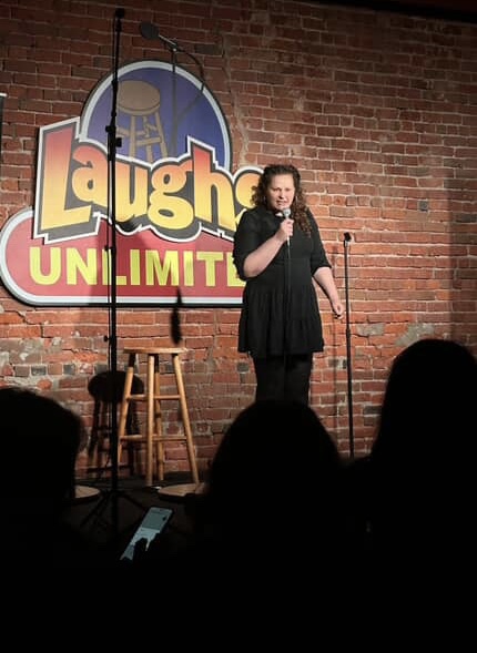 Female comedian performing on stage with a microphone, brick wall background, Laughs Unlimited sign, audience silhouettes.