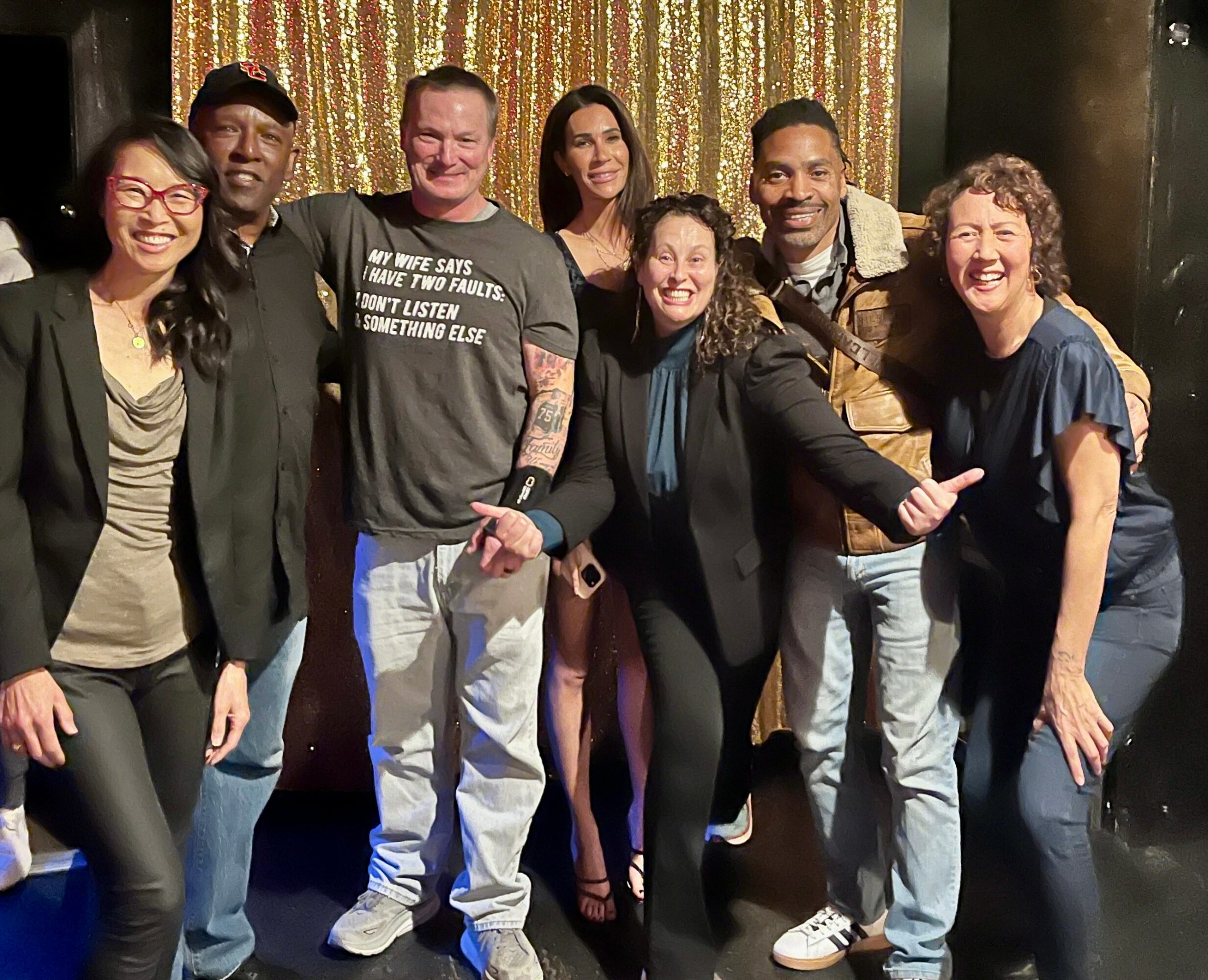 Group of seven diverse people smiling and posing together in front of a gold curtain backdrop.
