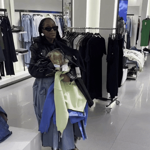 Person shopping in clothing store holding multiple jackets, wearing sunglasses and a black jacket, with racks of clothes behind.