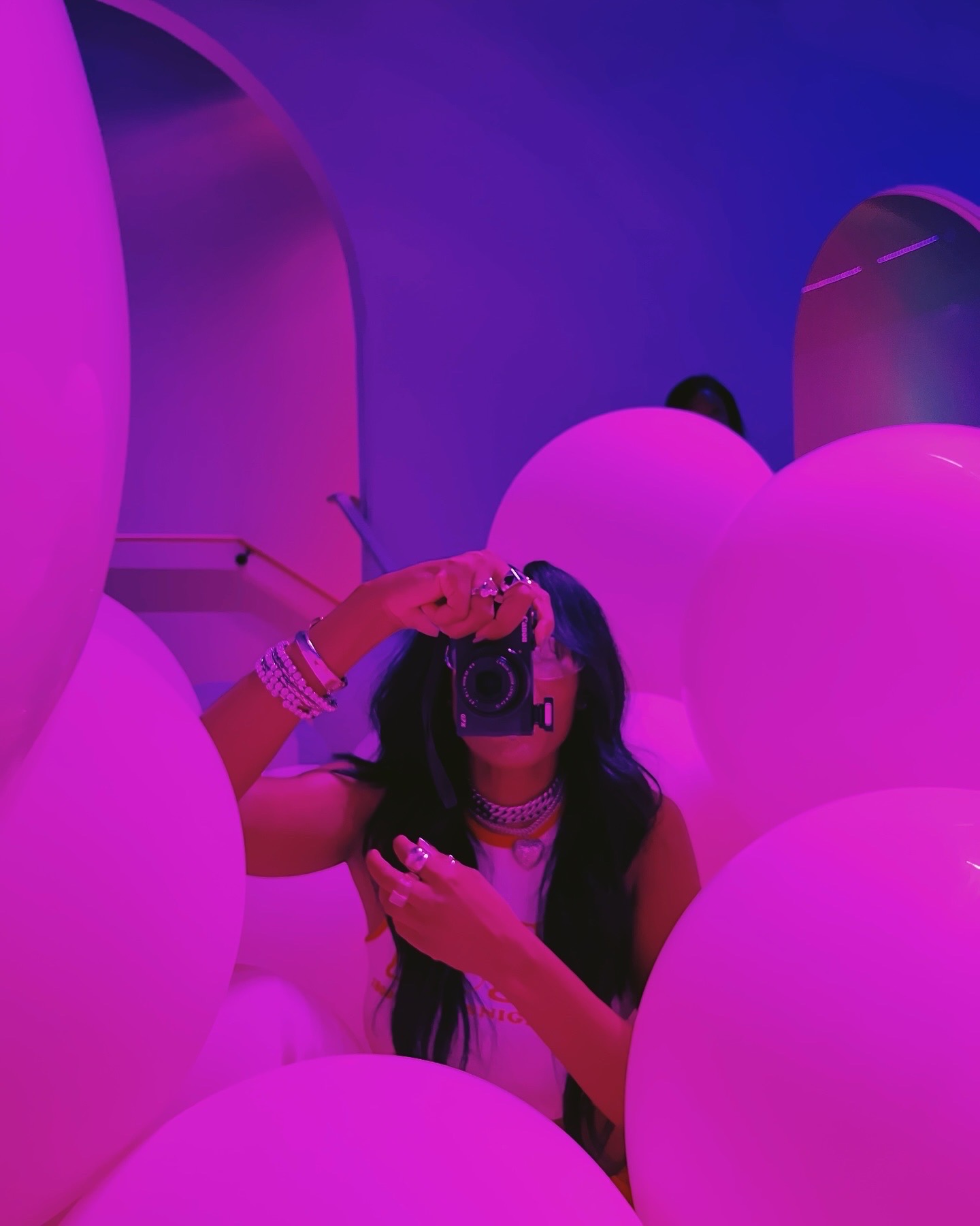 Person taking a photo with a camera surrounded by pink balloons and purple lighting.