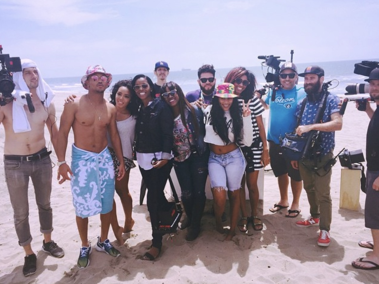 Group of people standing on a beach, some wearing swimsuits and casual clothing, with cameras and equipment nearby.