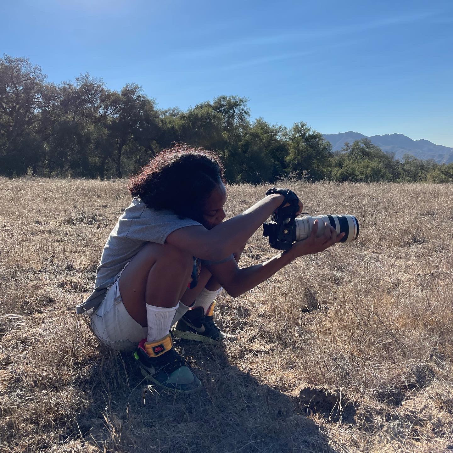Child crouching in a field, holding a camera, with trees and mountains in the background under a clear sky.