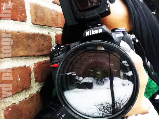 Person holding a Nikon camera with a large lens, standing near a brick wall, snow on the ground.