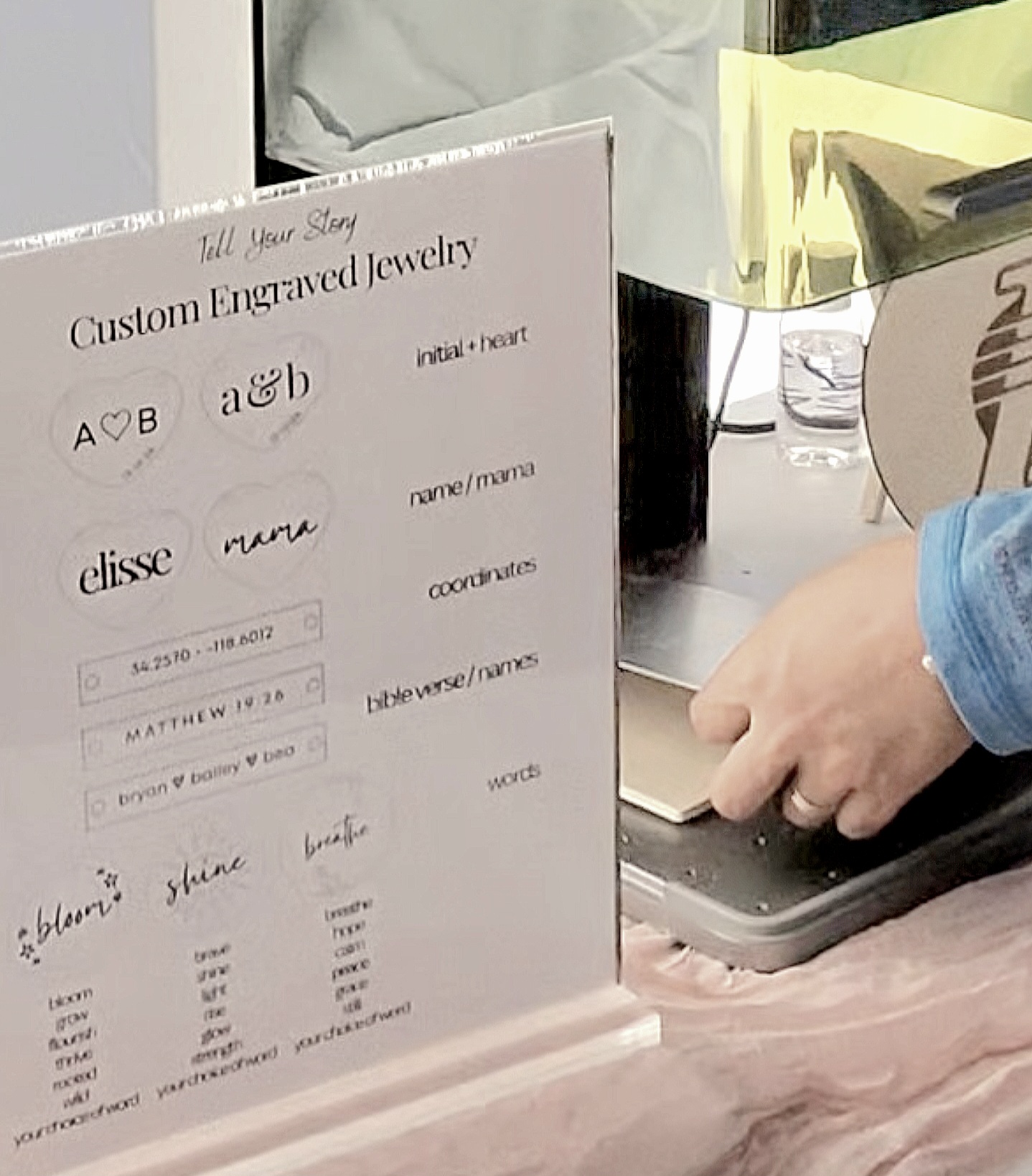 Sign displays custom engraved jewelry options with names, initials, and symbols, on a pink table with a person's hand nearby.