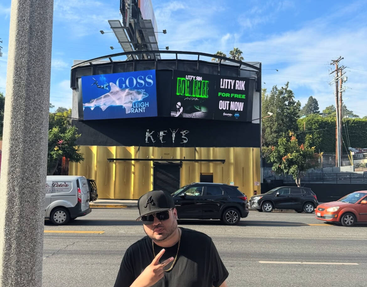 Young man wearing sunglasses and a black cap making a peace sign in front of a city street with cars and a billboard.