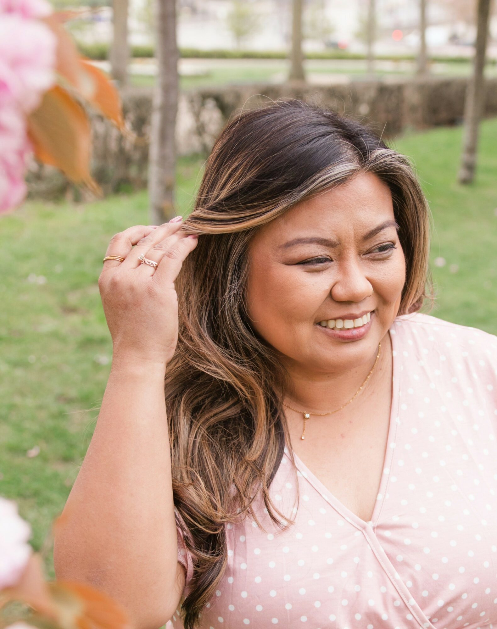 Woman with long hair smiling outdoors, touching her hair, in a park setting.